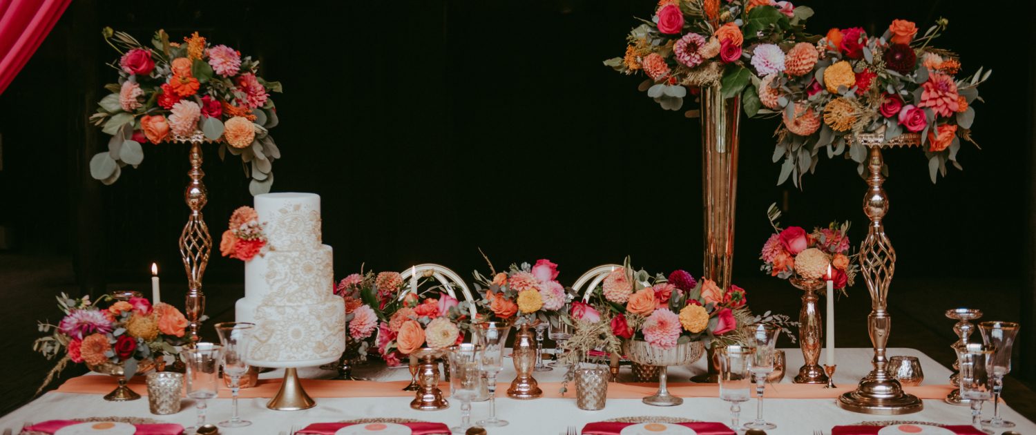 Bold fuchsia and orange tablescape designed with high and low floral arrangements, gold accented place settings and a white wedding cake.