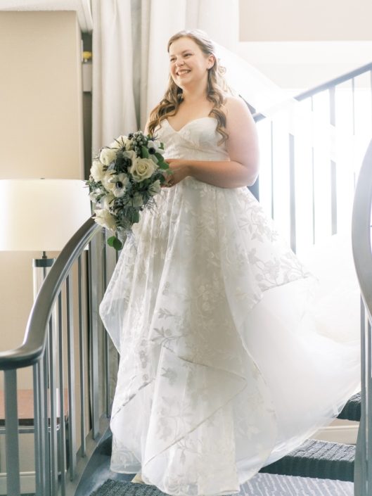 Bride walking down stairs with elegantly whimsical white and silver bouquet featuring roses, ranunculus, panda anemones