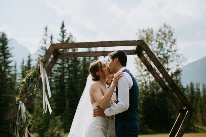 Bride and groom kissing at ceremony under a hexagon archway with greenery arrangement