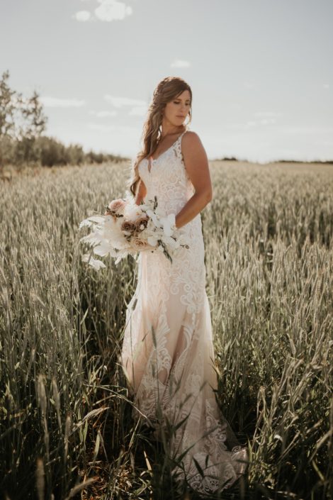 Bride in a field with rustic boho bridal bouquet featuring quicksand roses, amnesia roses, ranunculus, phalenopsis orchids, astilbe, bunny tail, bleached bracken fern and olive branches