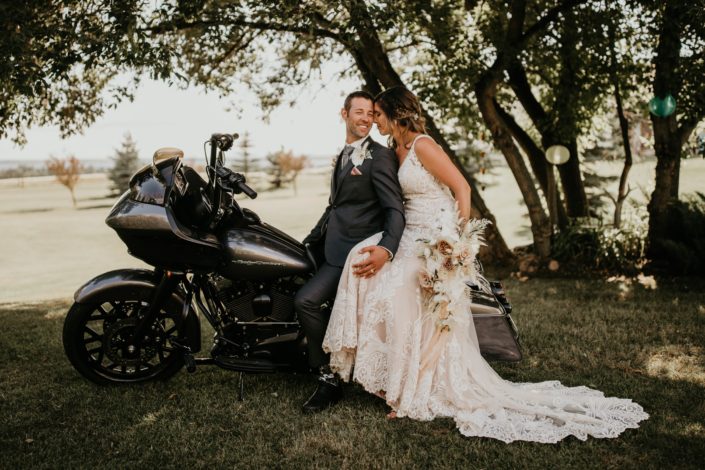 Bride and groom on a motorcycle with bridal bouquet featuring roses, ranunculus, phalenopsis orchids, bunny tail, bleached bracken fern, astilbe, olive branches and eucalyptus
