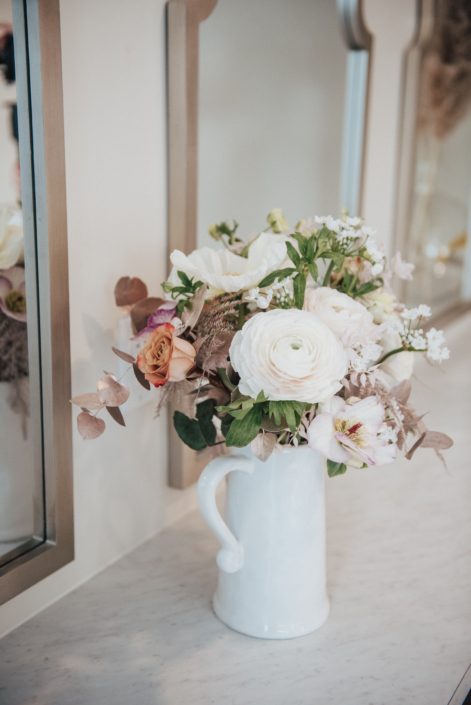Floral arrangement in a jug designed with roses, ranunculus, alyssum, and rose gold painted plumosa and eucaluptus greenery