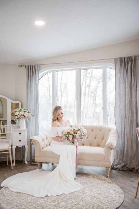 Bride in getting ready suite sitting on a sofa holding a pink and white bridal bouquet with trailing ribbons