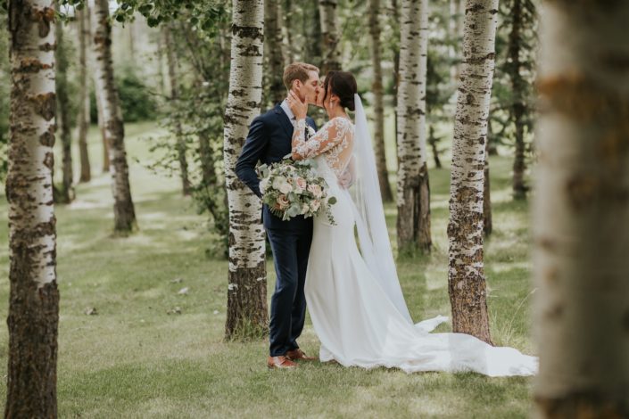 Bride and groom kissing amongst trees with bride's bouquet