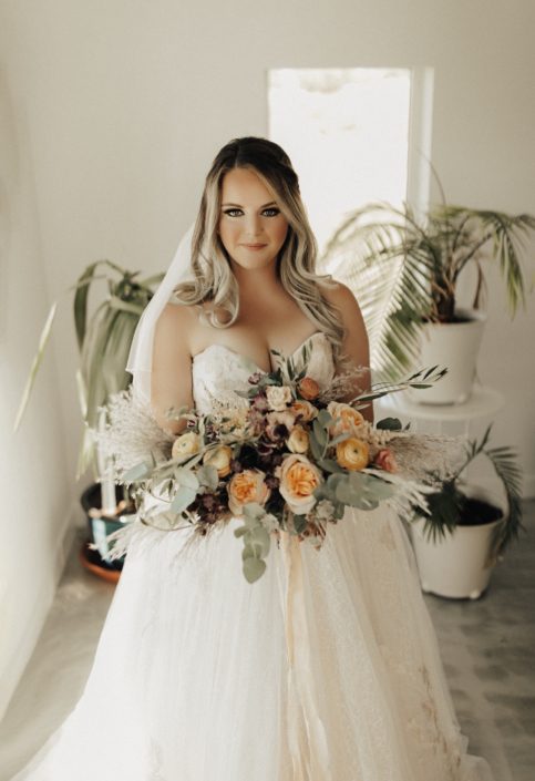 Bride with boho bridal bouquet designed in peach and burgundy tones with flowers such as Juliet Garden roses, dahlias, ranunculus, pampas grass and olive branches