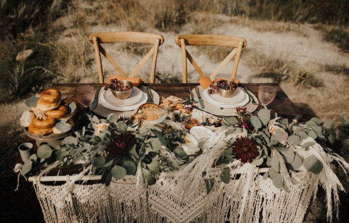 Sweetheart table with charcuterie, donuts, flowers and wooden chairs; boho