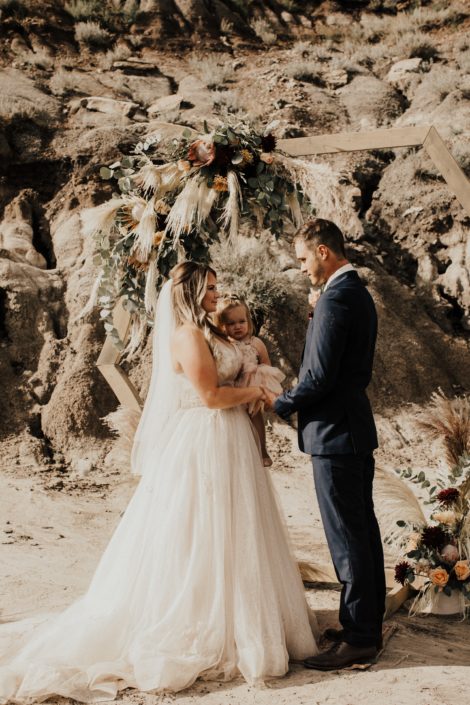 Bride and groom in front of outdoor ceremony wooden archway covered with boho floral arrangements