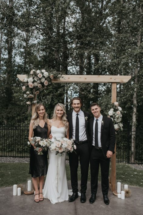 Bride and bridesmaids with pastel bouquets under a wooden archway decorated with floral arrangements of roses, poppies, peonies, ranunculus and greenery