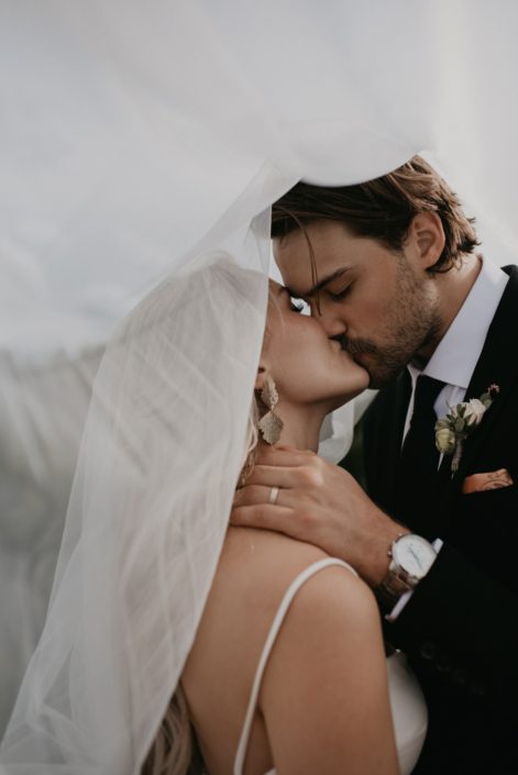 Bride and groom kissing under veil with groom's boutonniere designed with white spray roses