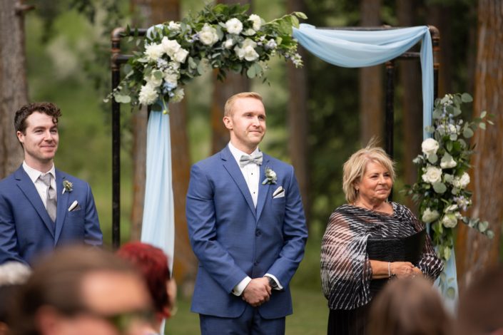 Groom wearing blue suit and boutonniere under archway covered with blue and white flowers such as delphinium, eryngium, ranunculus and roses