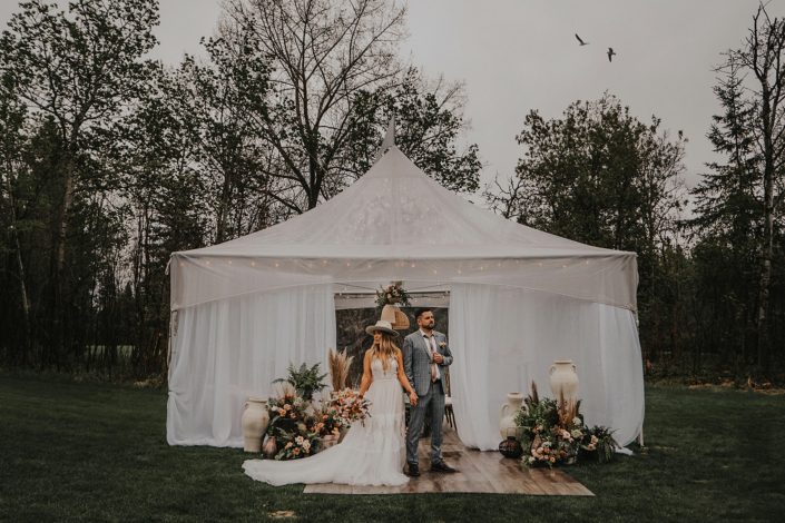 Modern boho couple standing in front of tent with rust/terracotta toned flowers