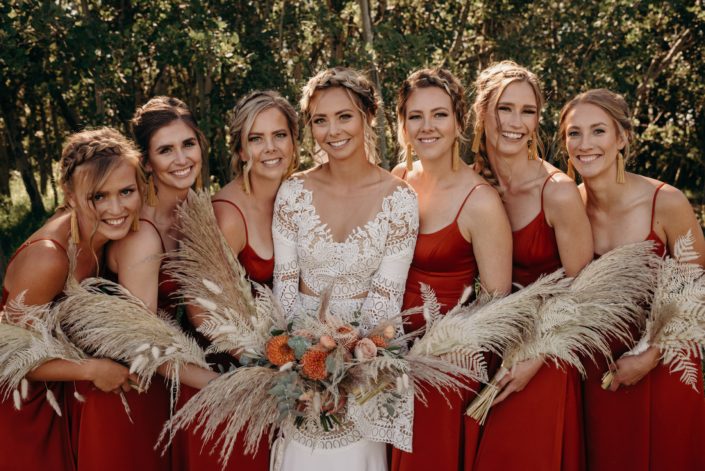 Orange boho - Bride and bridesmaids with bouquets; bridal bouquet designed with orange dahlias, ranunculus, cappuccino roses, bleached bracken fern, bunny tail, eucalyptus, olive branches and pampas grass; bridesmaid bouquets designed with pampas grass, bleached bracken fern and bunny tail