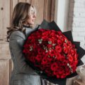 A model holds an impressively large hand tied bouquet of 100 rich, velvety red roses accented with some greenery and finished off with black Korean paper wrap.