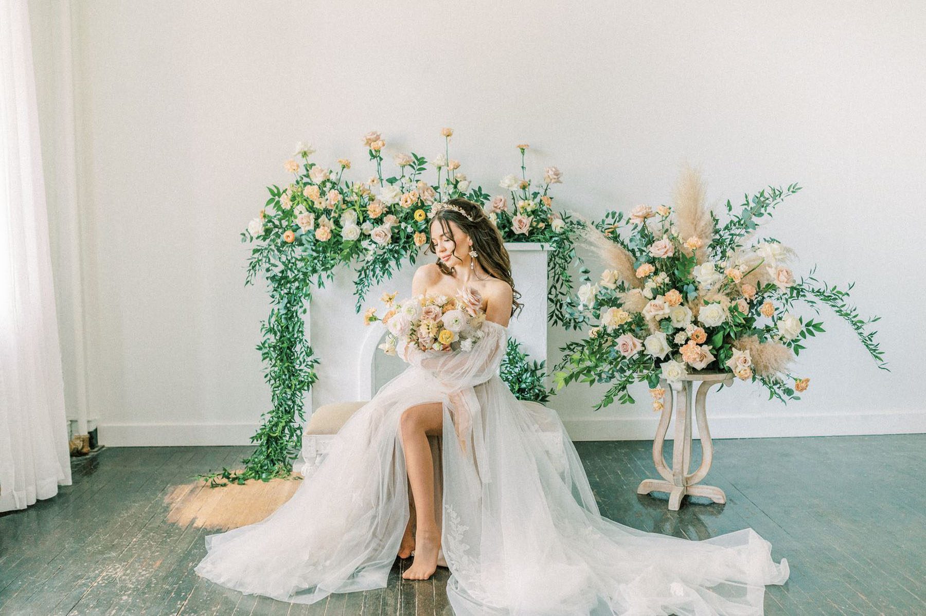 Bride with light yellow, pink and white bouquet in front of a white fireplace covered in draping wedding florals and a side table with large arrangement