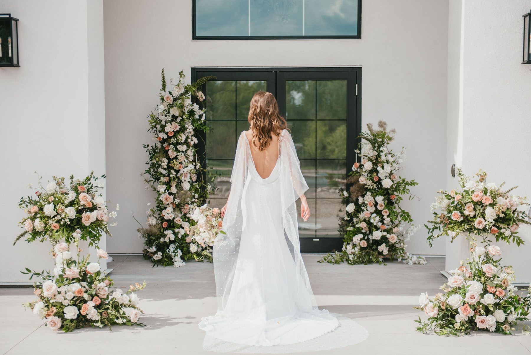 Bride standing in front of a glass door surrounded by peach, blush, white floral arrangements