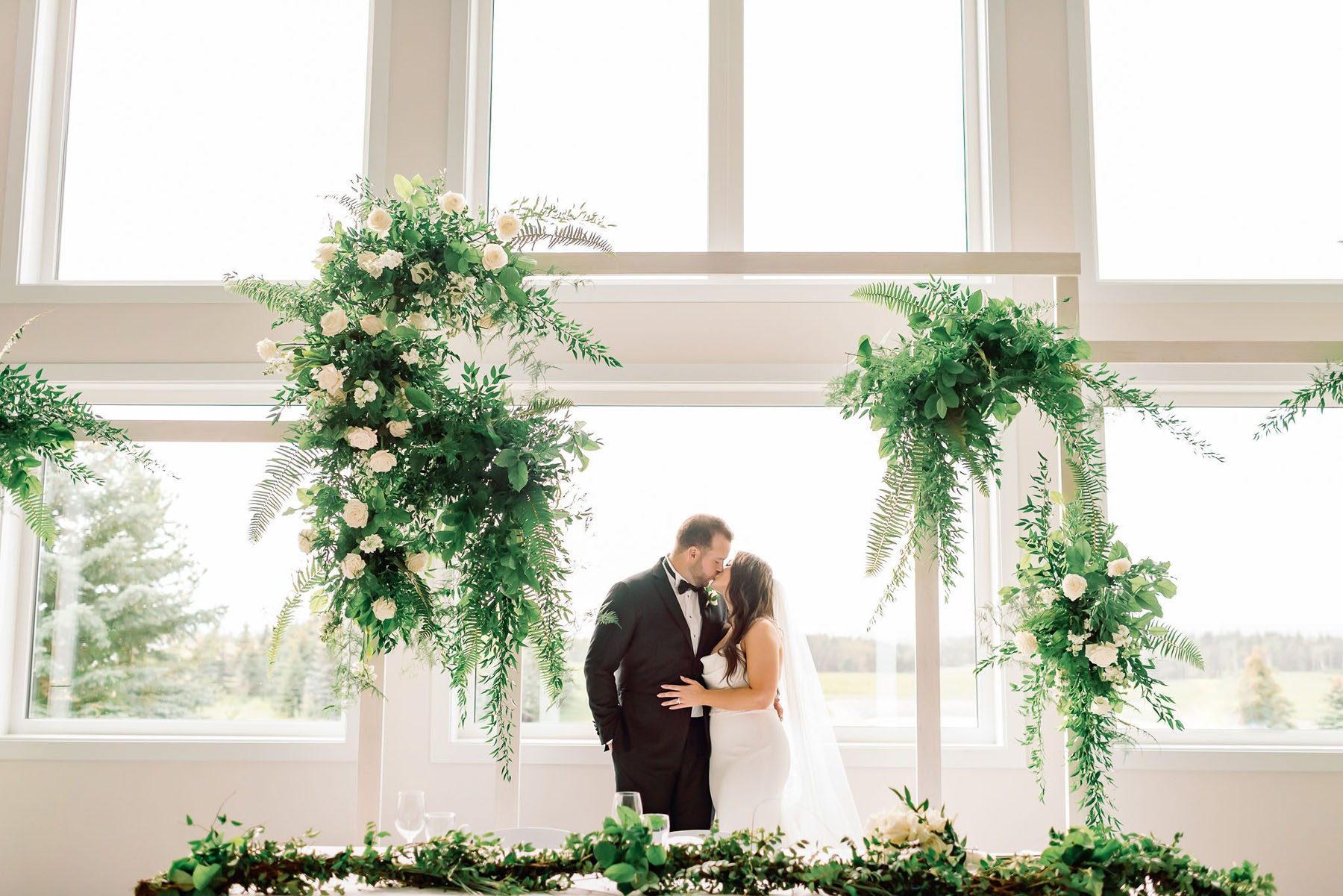 Bride and groom kissing behind a head table decorated with branches and greenery. Multiple large fern, leaves and blush flower arrangements frame them.