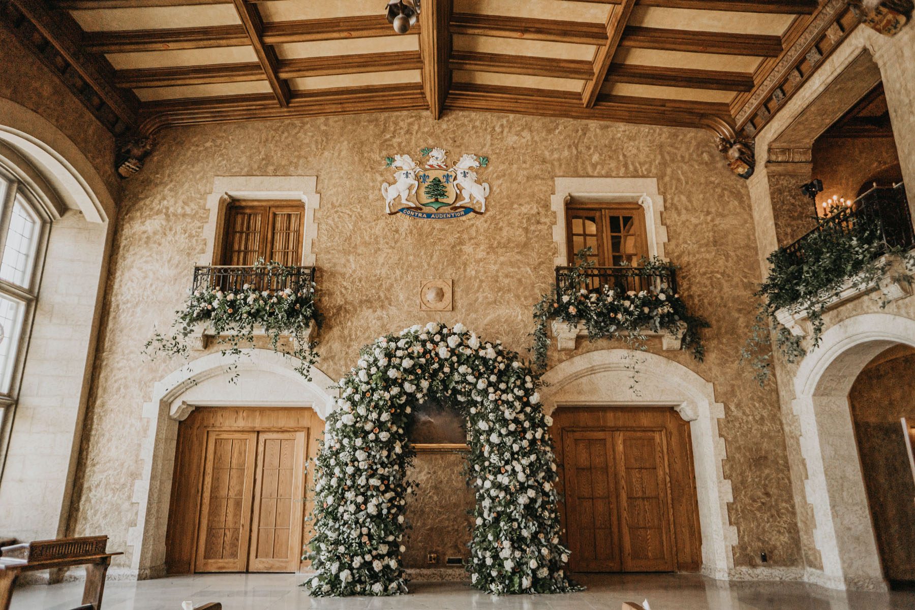 Grand wedding arch with blush white and greenery placed between 2 large doors below window balconies draped with florals