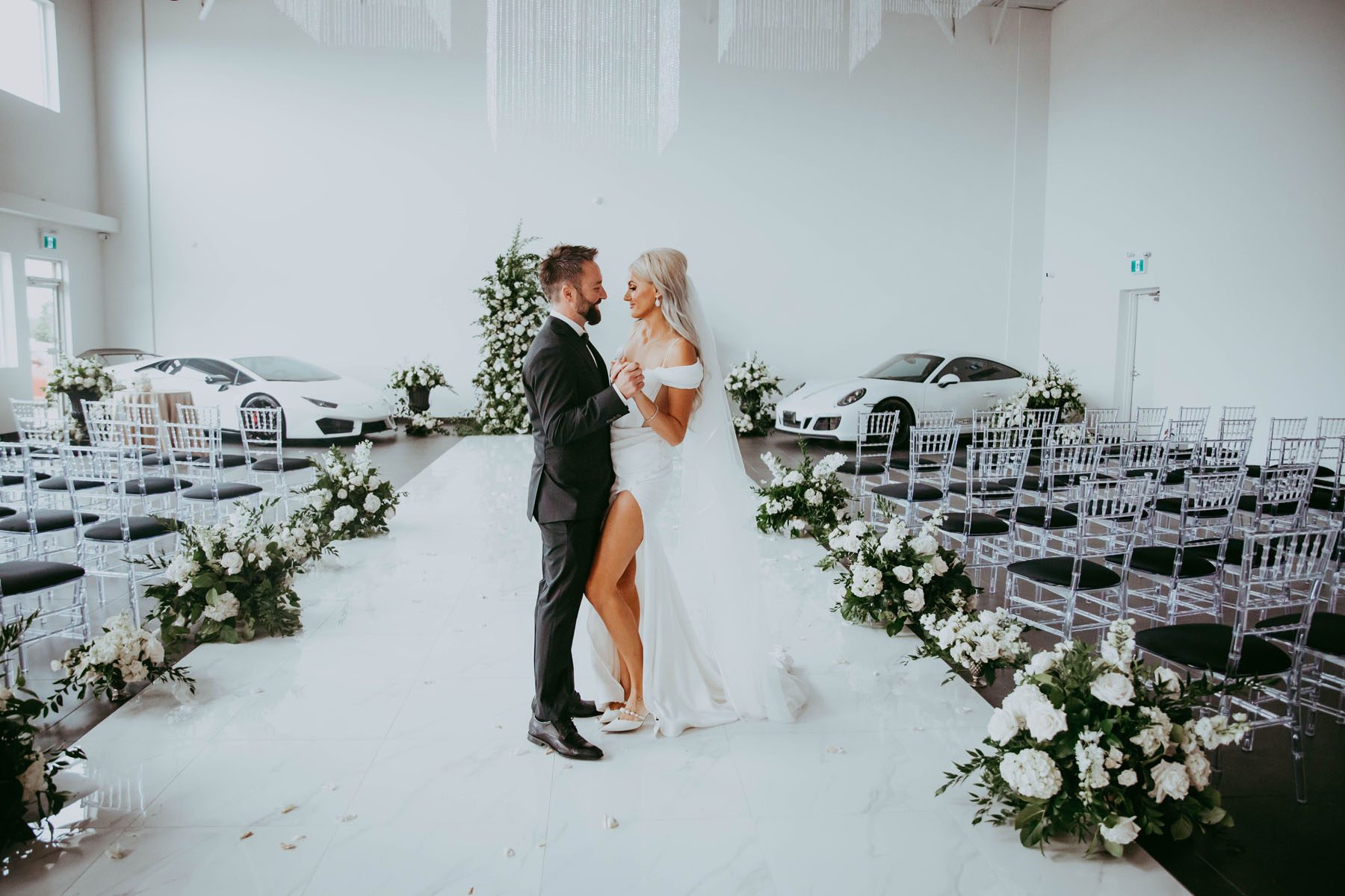 Bride and groom standing among white and green floral row markers with 2 white sports cars and a floral arch