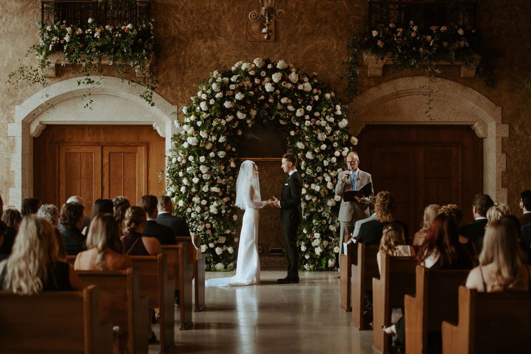 Bride and groom standing under a grand floral arch with white, blush and greenery.