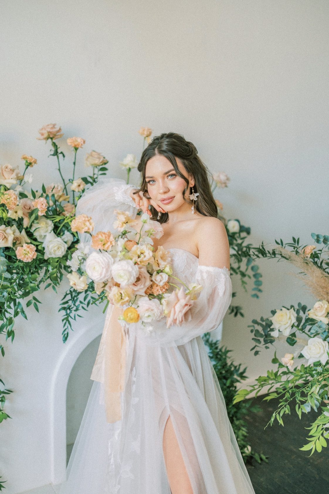 Our model standing in front of a faux fireplace with a large floral arrangement along the mantel, holding up her peach and blush bridal bouquet