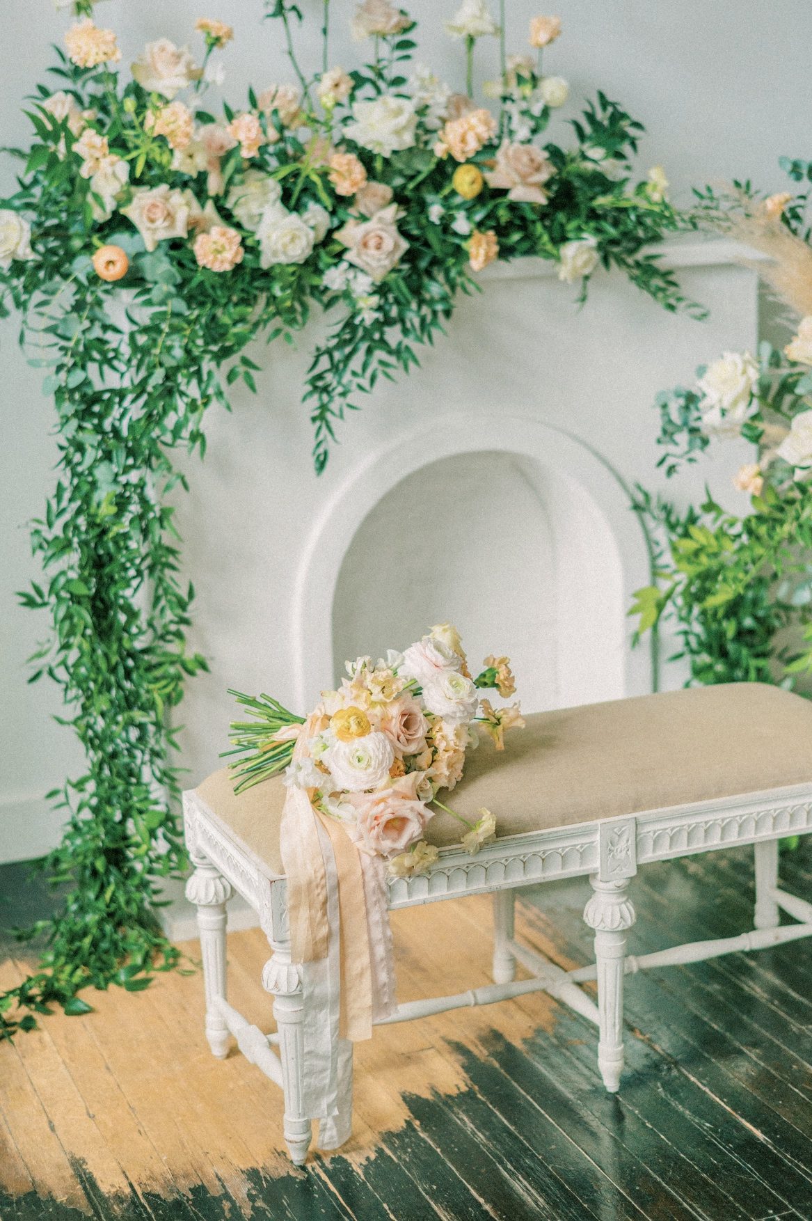 A faux fireplace with a large floral arrangement in peaches and blush over the mantel with lots of greenery draping over the side. In front a white bench, with the bridal bouquet in matching flowers is set on its side
