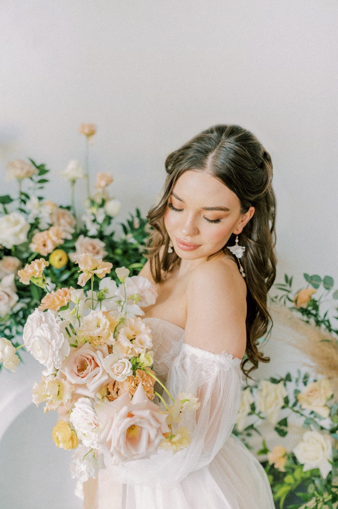 A Calyx Floral Design model stands in front of large peach floral arrangements, holding up her bridal bouquet and gazing down at it