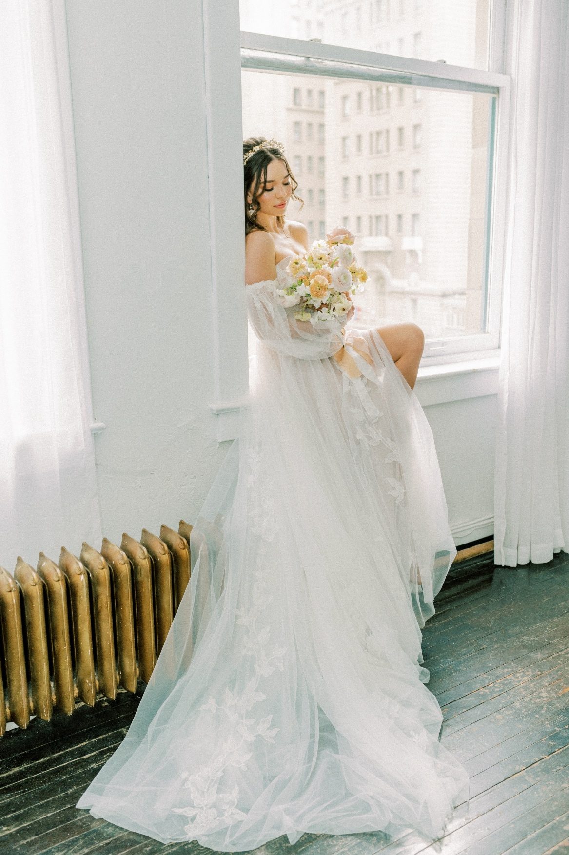 Our bride sits inside a window well, with her dress elegantly draping down. She looks down at her peach bridal bouquet