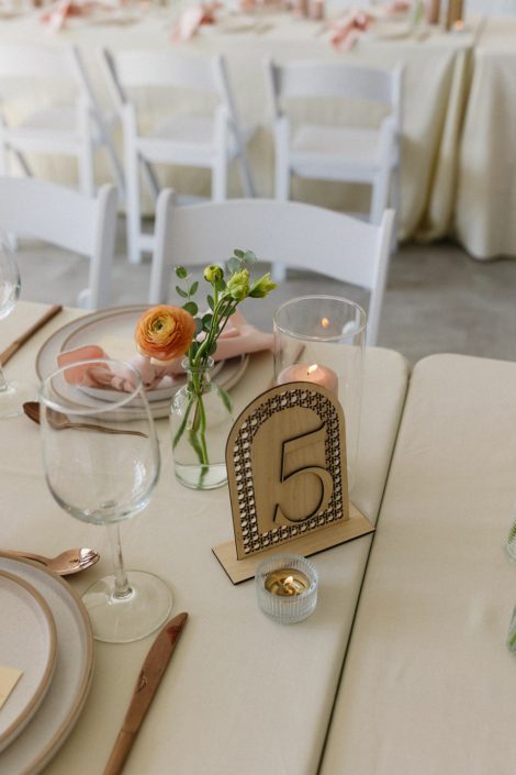 Detail photo of a tablescape with a bud vase centerpiece with a couple of blooms and a sprig of greenery, a wooden table number and a vase with a pillar candle.