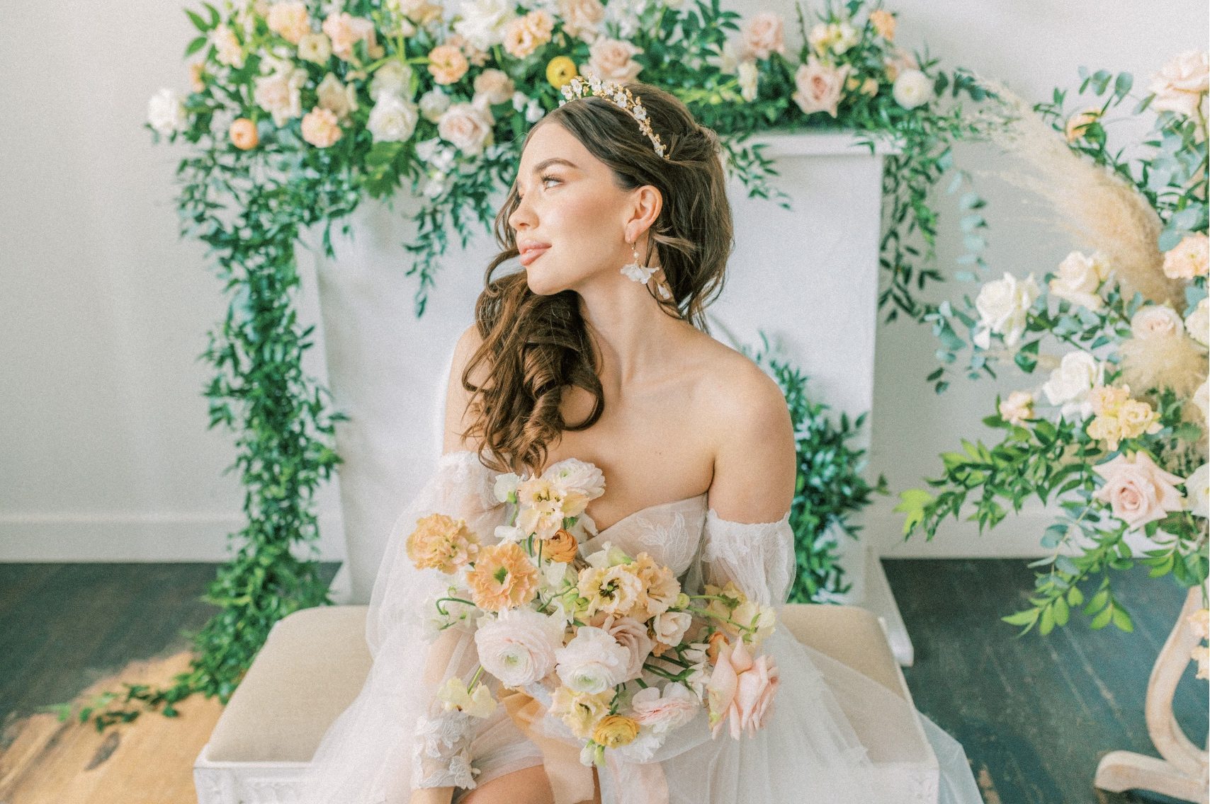 Calyx Floral Design model sits on a bench in front of a faux fireplace decked out in a large peach and green floral arrangement as she holds her bouquet and gazes out the window.