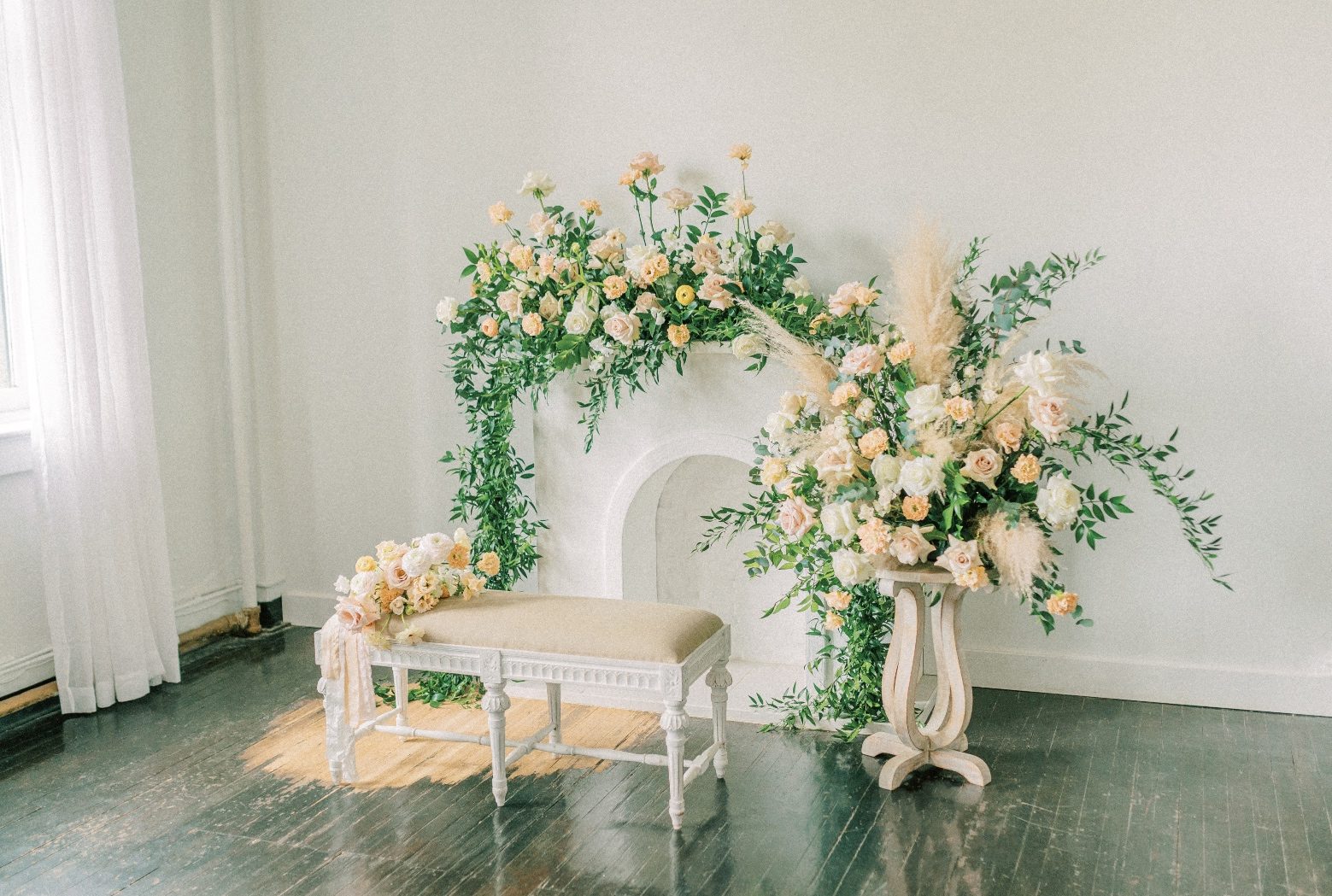 A shot capturing all of the Calyx Floral Design flowers for the photo shoot, including a large arrangement over the mantel of the faux fireplace, a large arrangement on top of a stand and a bench with the bridal bouquet set on it's side.