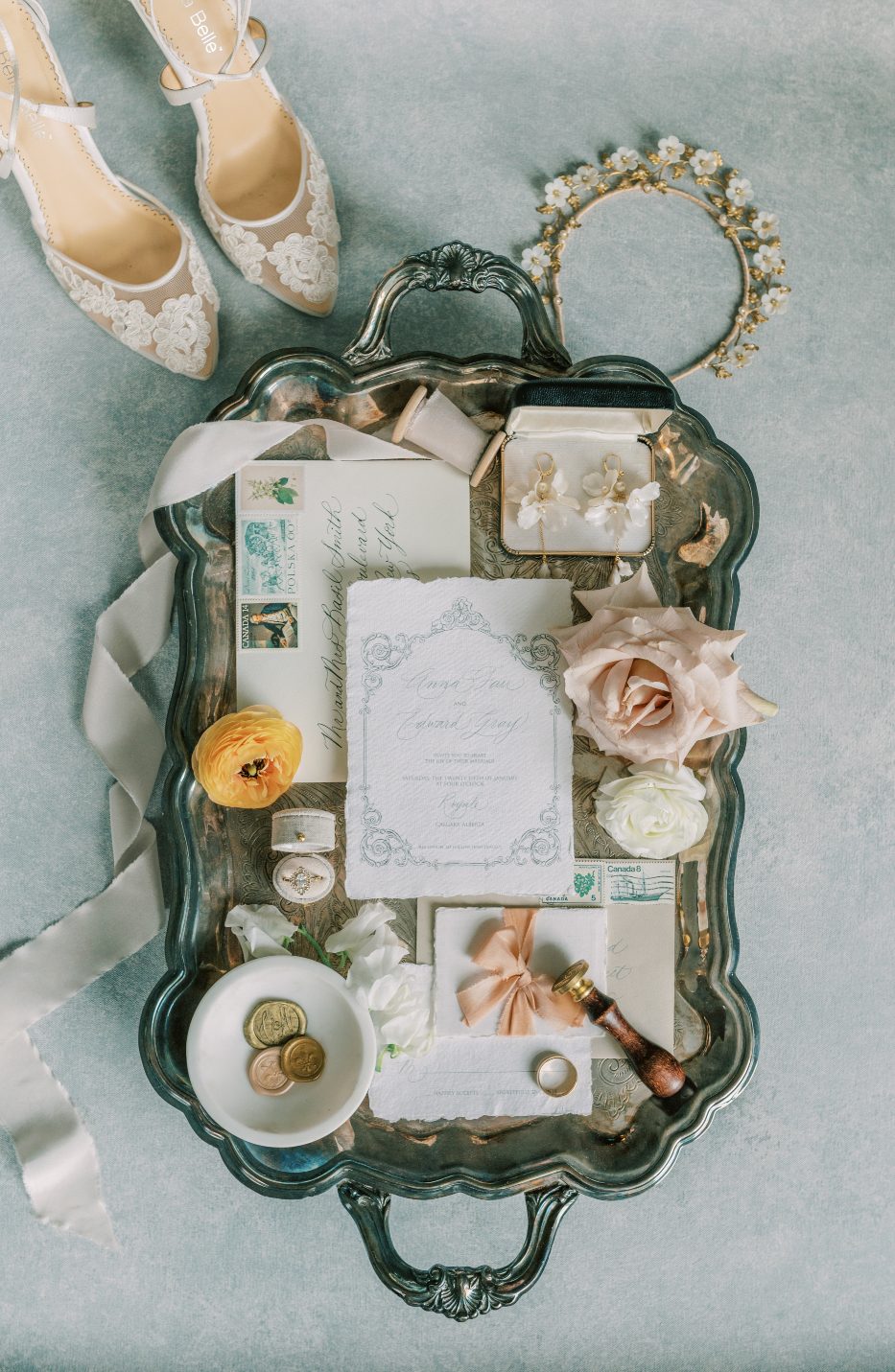 A flat lay shot of many detail pieces for the photoshoot including the wedding invitation, wax seals, wedding ring, jewelry and shoes accented with pretty blooms