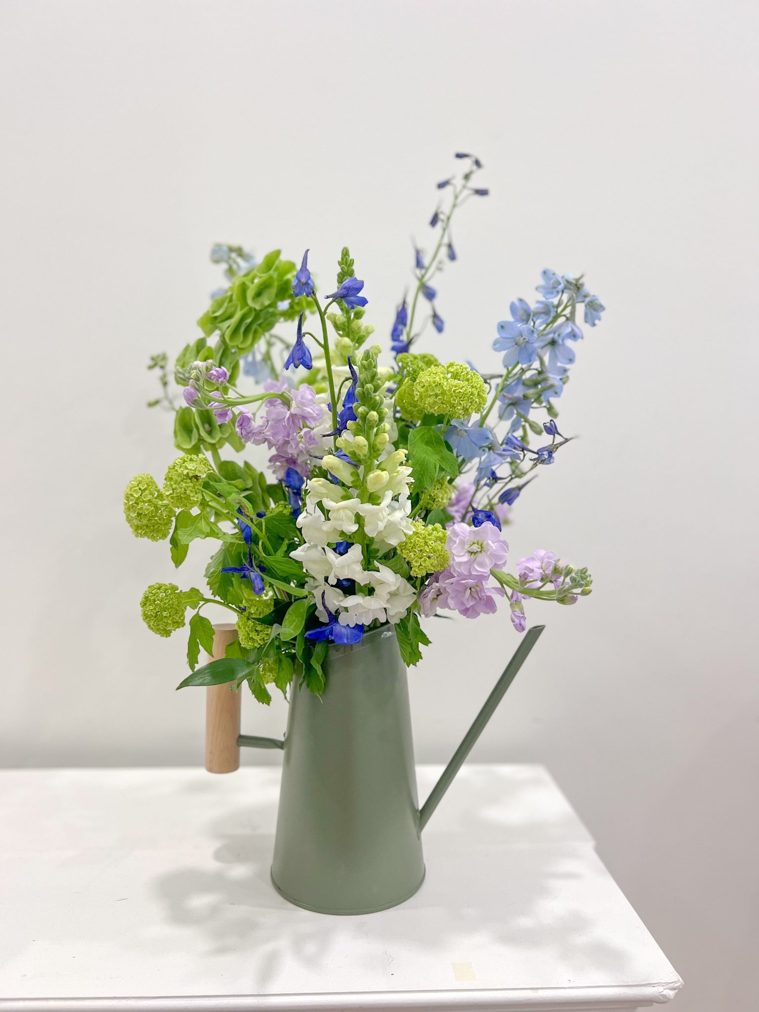 An upright garden arrangement in greens, blues and purples arranged into an olive green watering can