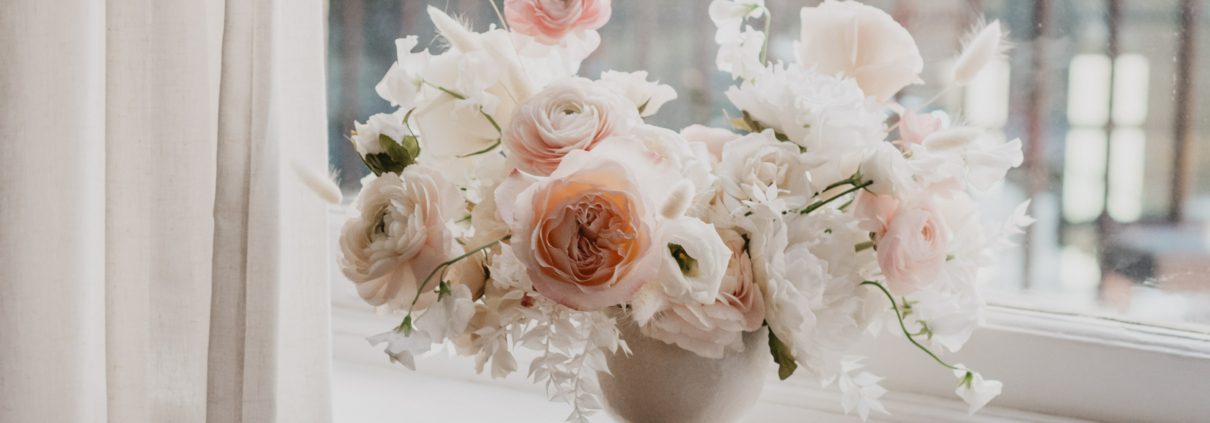 A pretty arrangement of white and blush in a white pedestal vase sits on a window sill.