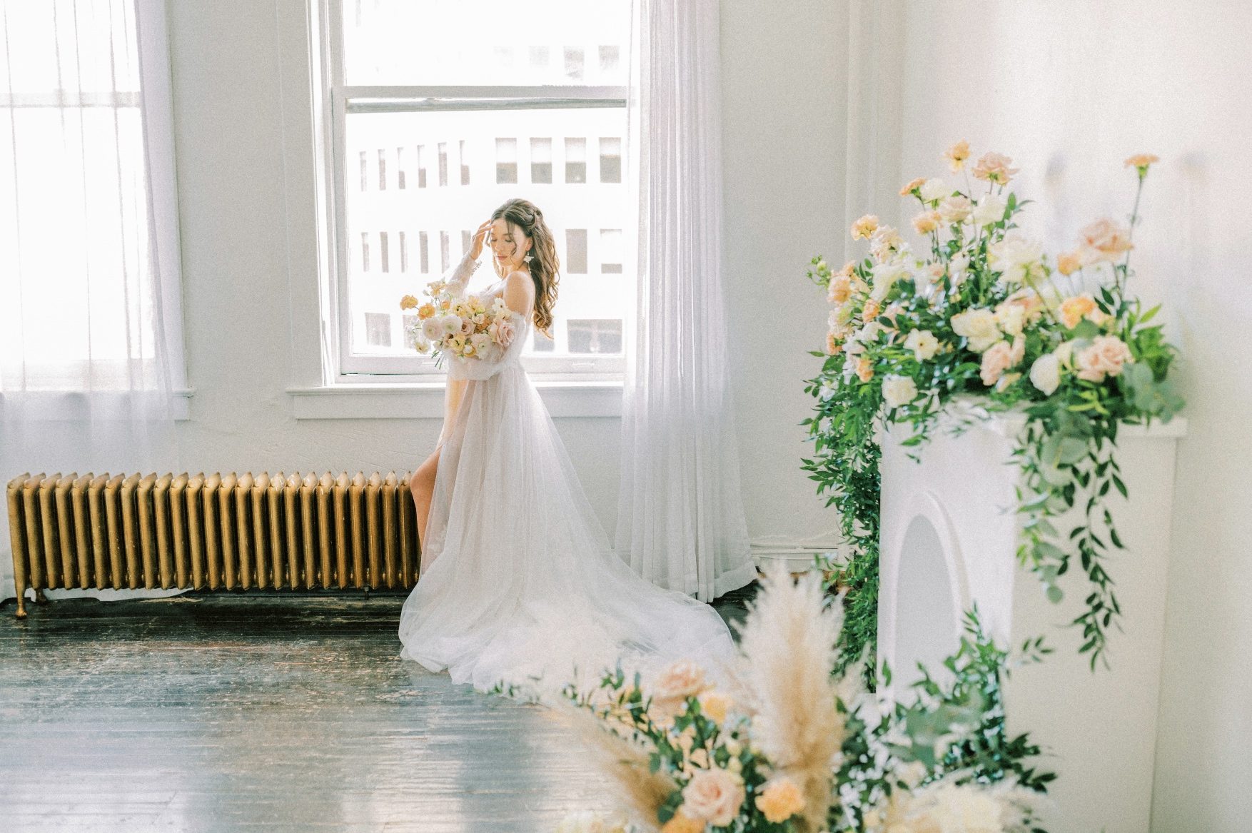 A Calyx Floral Design model stands in front of a window streaming with natural light. Her beautiful gown pools around her feet and she holds a peach and blush bouquet