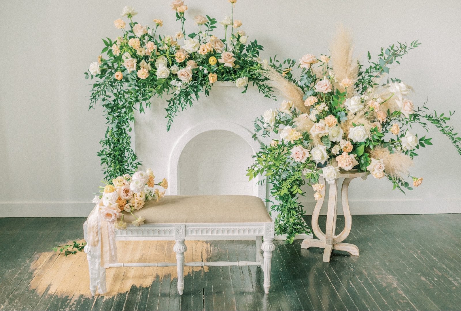 All of the floral pieces for the photoshoot - the bridal bouquet set on a bench in front of a faux fireplace with a large floral arrangement along the mantel and beside it a big floral arrangmeent in coordinating colours on top of a decorative stand.