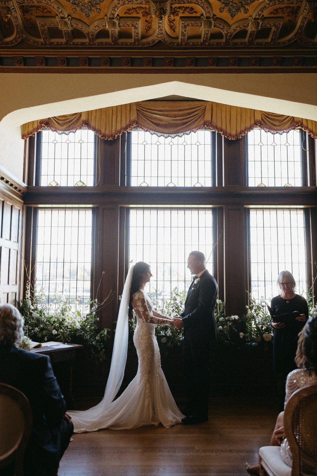 Stephanie and Tanner stand clasping hands in front of the large windows and floral arrangements, ready to say 'I do'.