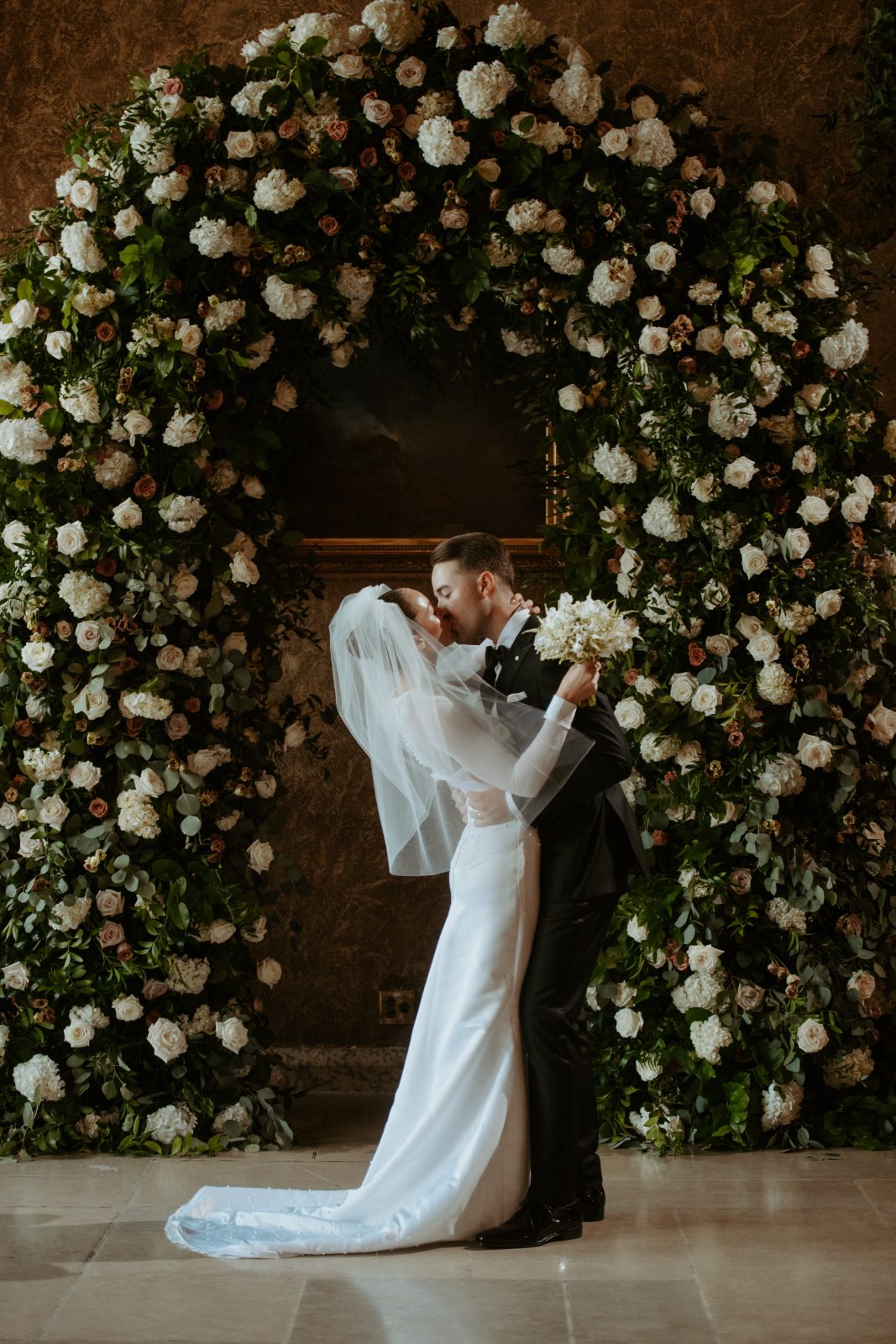 A Calyx couple embraced in a kiss for their 'I do's' in front of a large floral arch