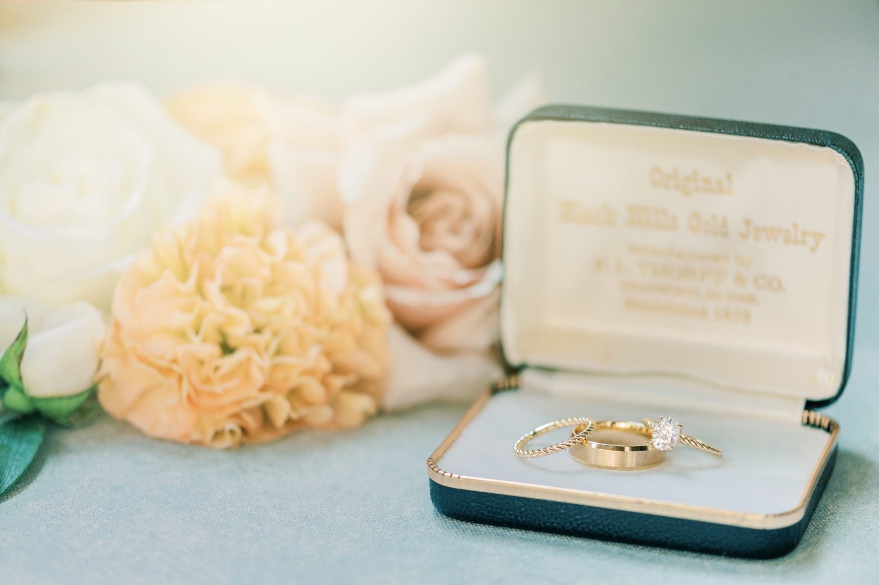 A second set of wedding ring and bands in a decorative jewelry box with peach and blush blooms in the background