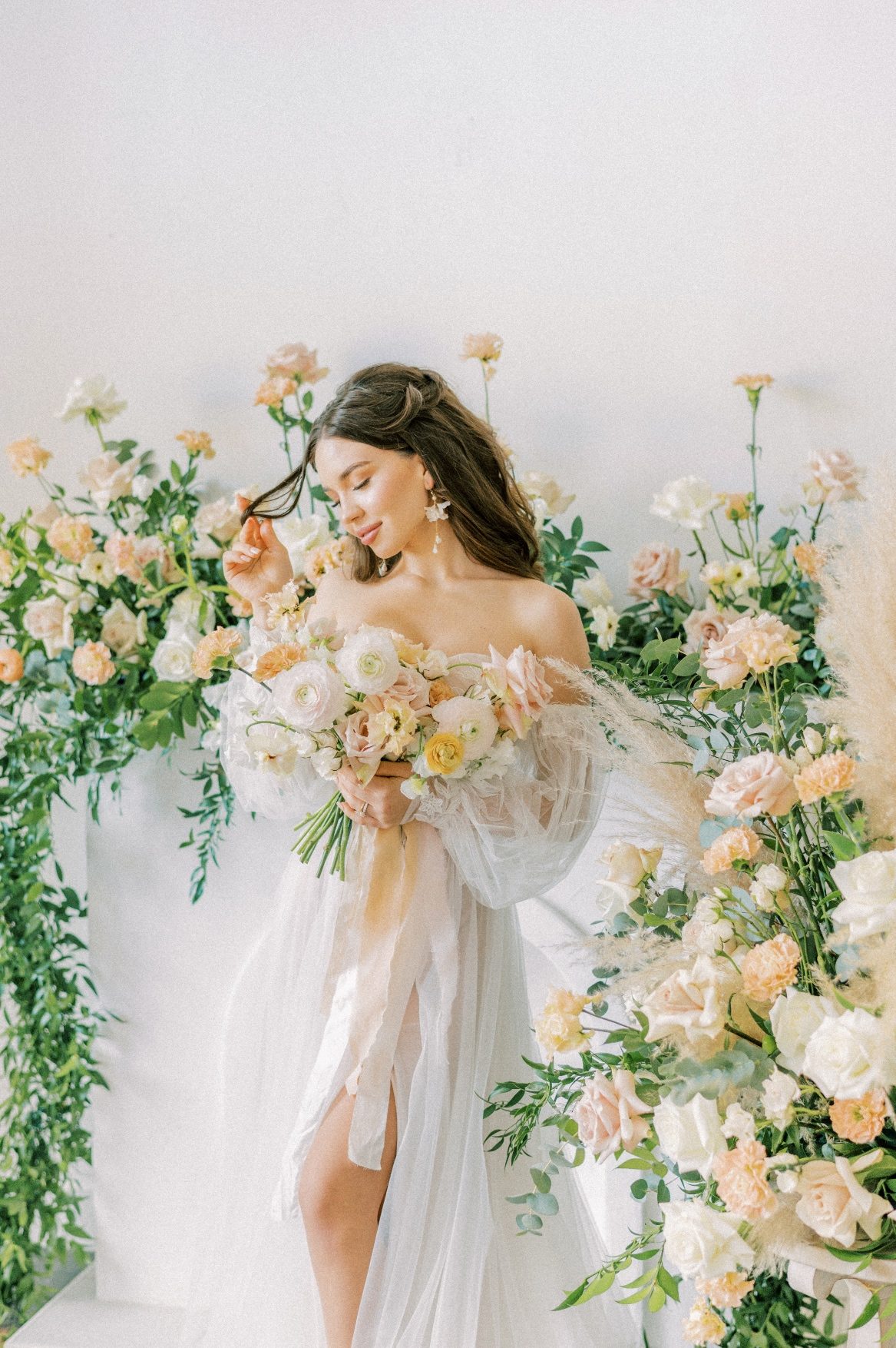 A Calyx Floral Design model twirls her hair around her finger with one hand, and holds her peach and blush bouquet in the other, with floral arrangements surrounding her.