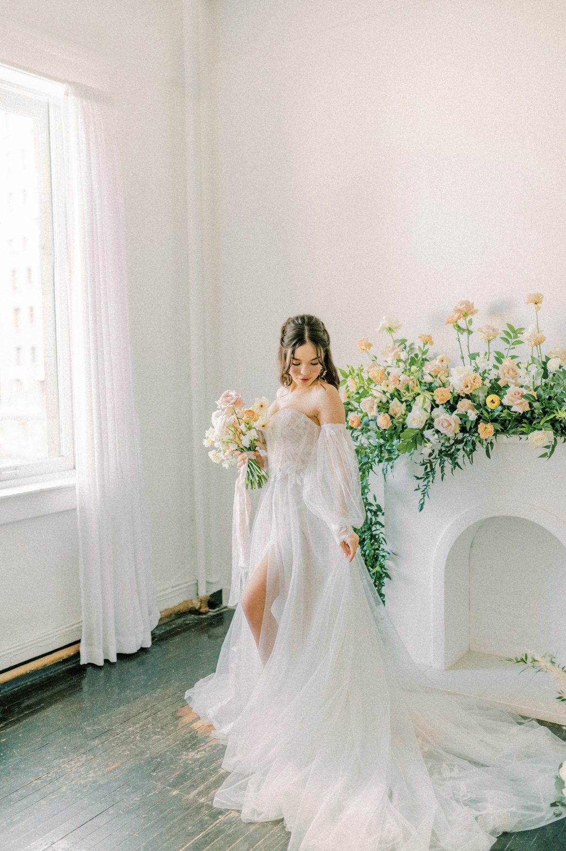 Our model stands in front of the beautiful mantel arrangement, holding her peach and blush bouquet looking down at the draping fabric of her bridal gown