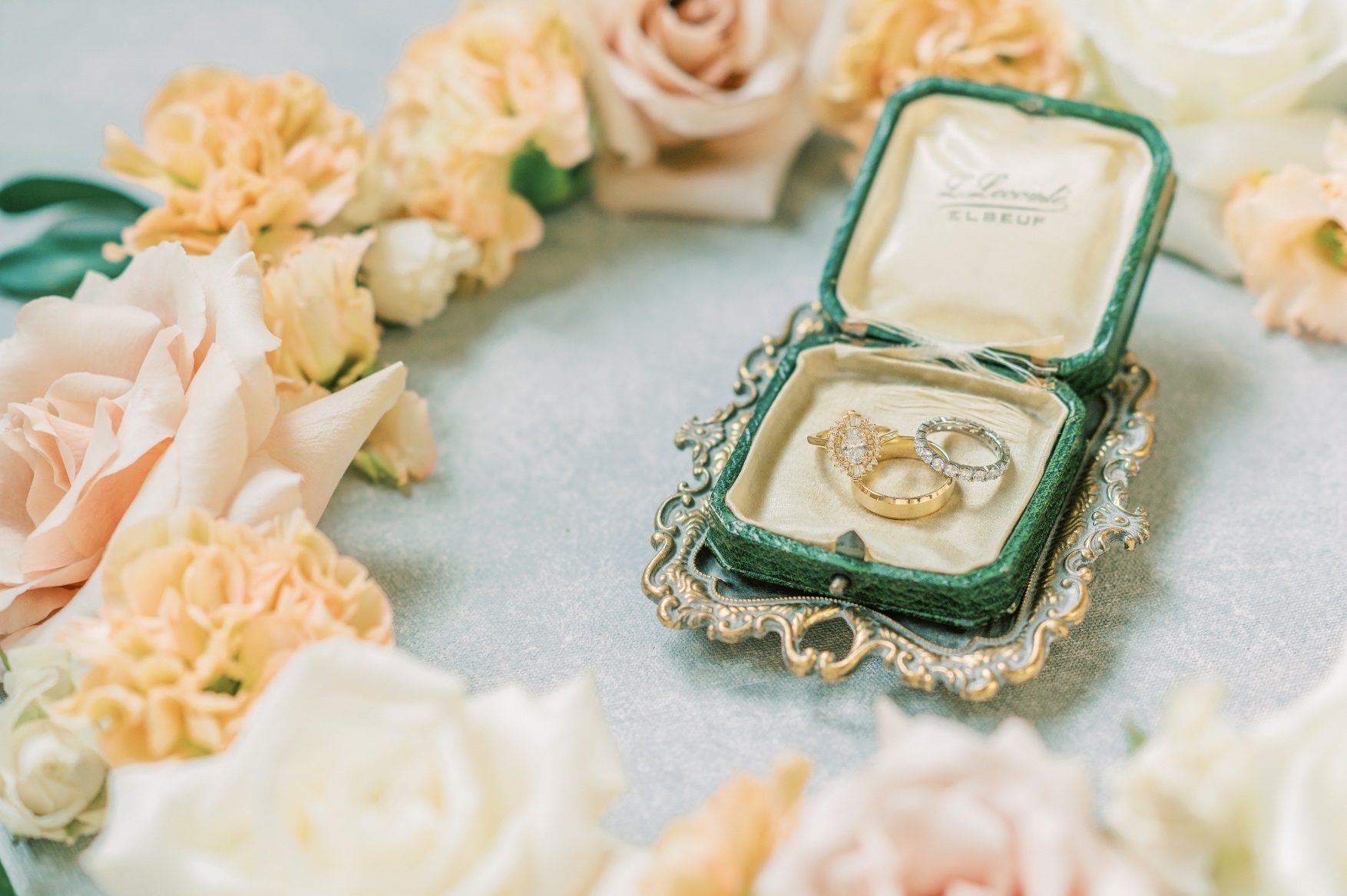 A pretty green jewelry box showcasing the brides ring and the wedding bands with a ring of flowers surrounding them.