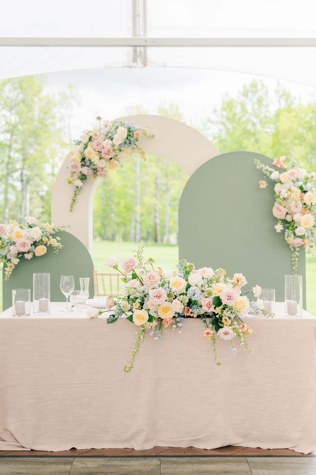 A mod backdrop with two sagey green curved pieces and one white curved piece adorned with pastel floral arrangements. A sweetheart table sits in front of it, also with a beautiful pastel centrepiece.