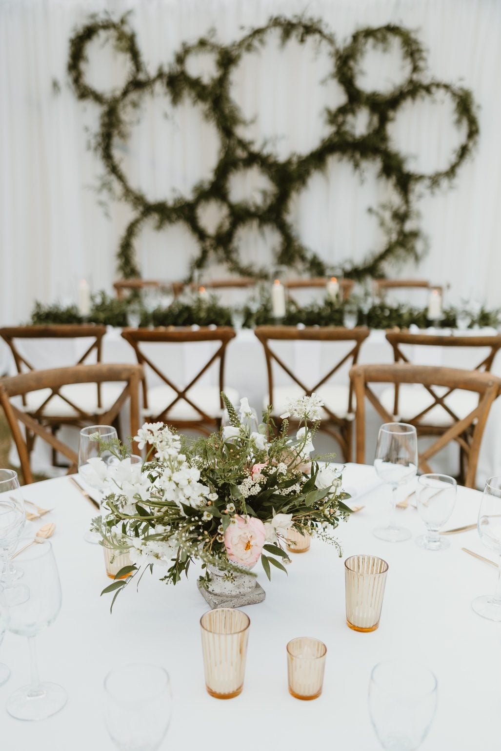 Wall Accent + Centrepiece A view of one of the round tables with a foral centrepiece with the greenery hoop wall accent in the background.