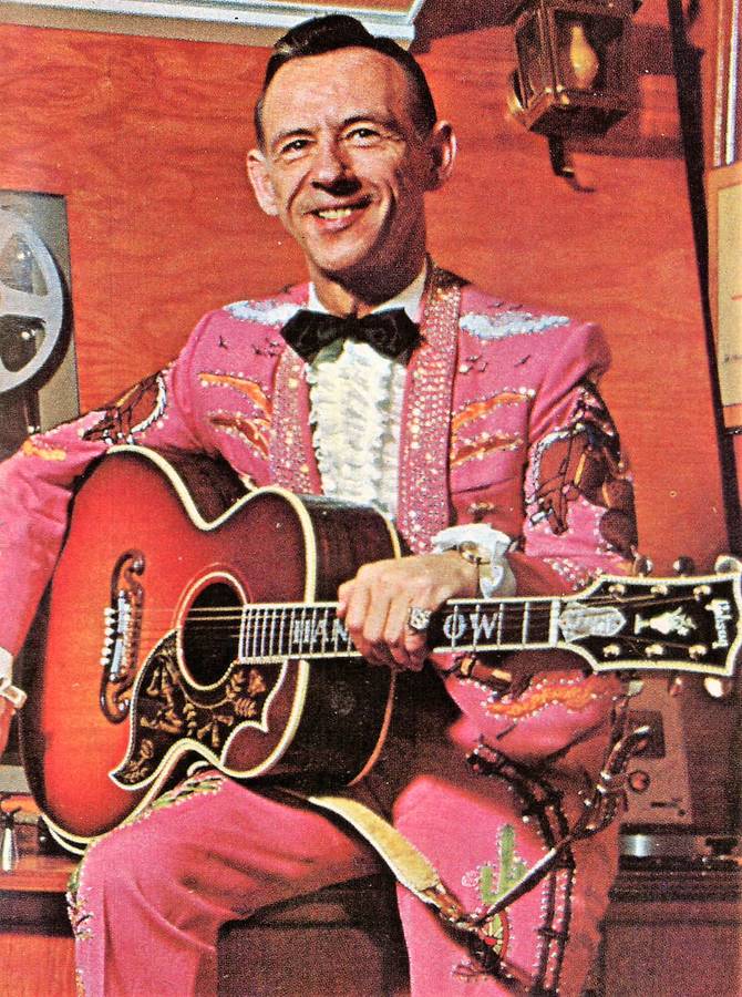 An inspiration of Hank Snow in a pink suit and black tie holding his guitar.