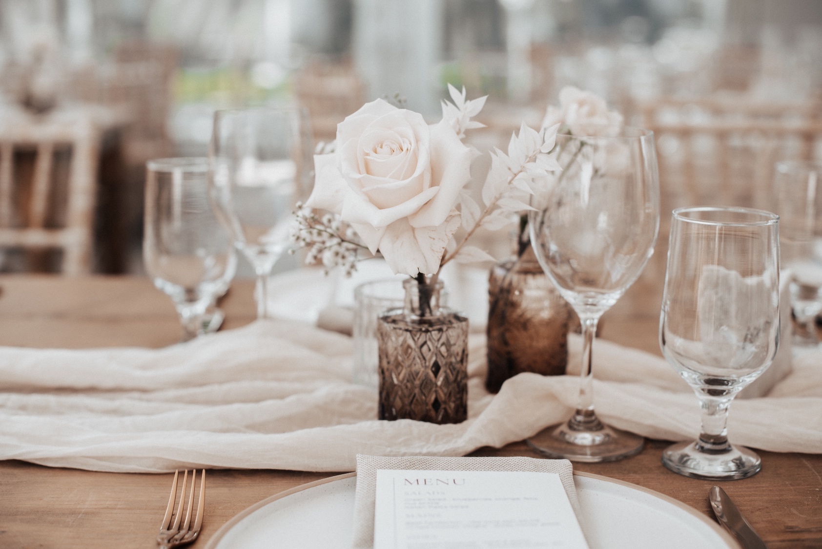 Budvase Centerpieces A grouping of bud vases made out of light brown glass, featuring white roses, bleached ruscus, baby's breath and ranunculus sitting in the middle of a soft gauzy table runner