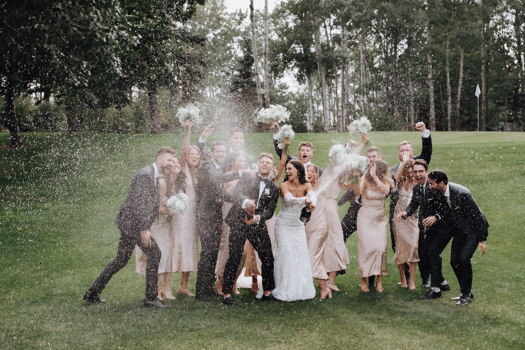Champagne Blast The couple and their wedding party are all standing closely together, clearly excited and celebrating while the bride and groom are spraying champagne everywhere.