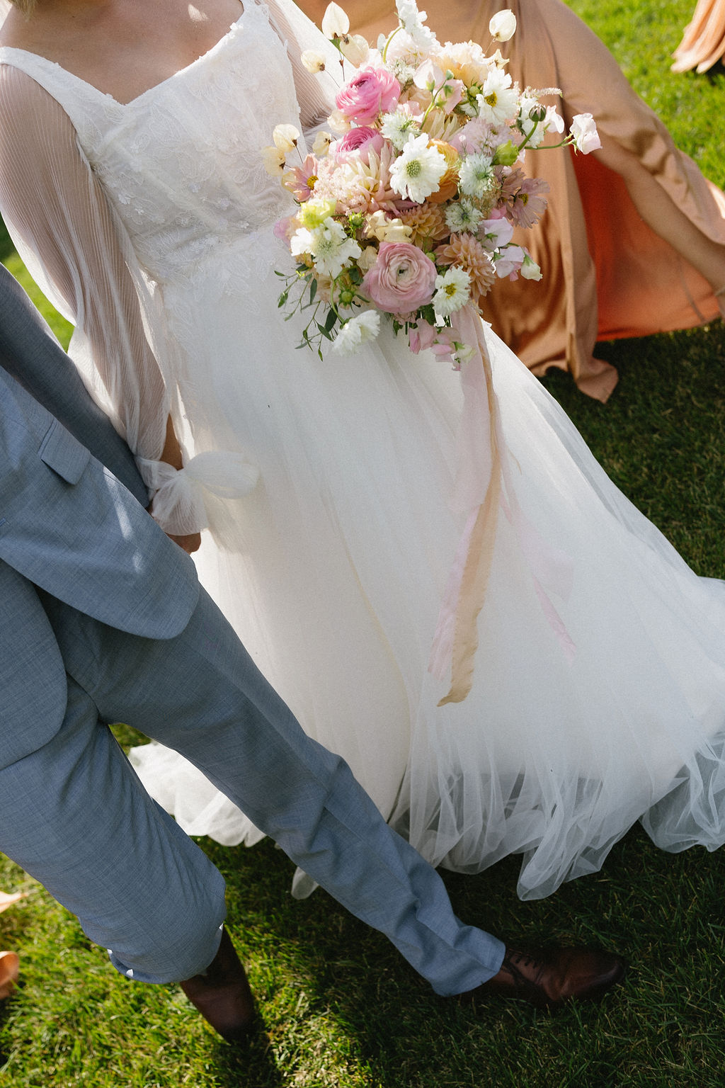 Happy Calyx Couple walking hand in hand, with the beautiful, pastel bridal bouquet with trailing blush and peach ribbons.