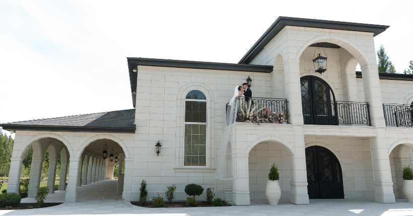 The breath taking scenery of Central Alberta's hottest new venue, Archway Manor. The bride and groom are standing at one of the beautiful balconies, with her long veil trailing over the railing.