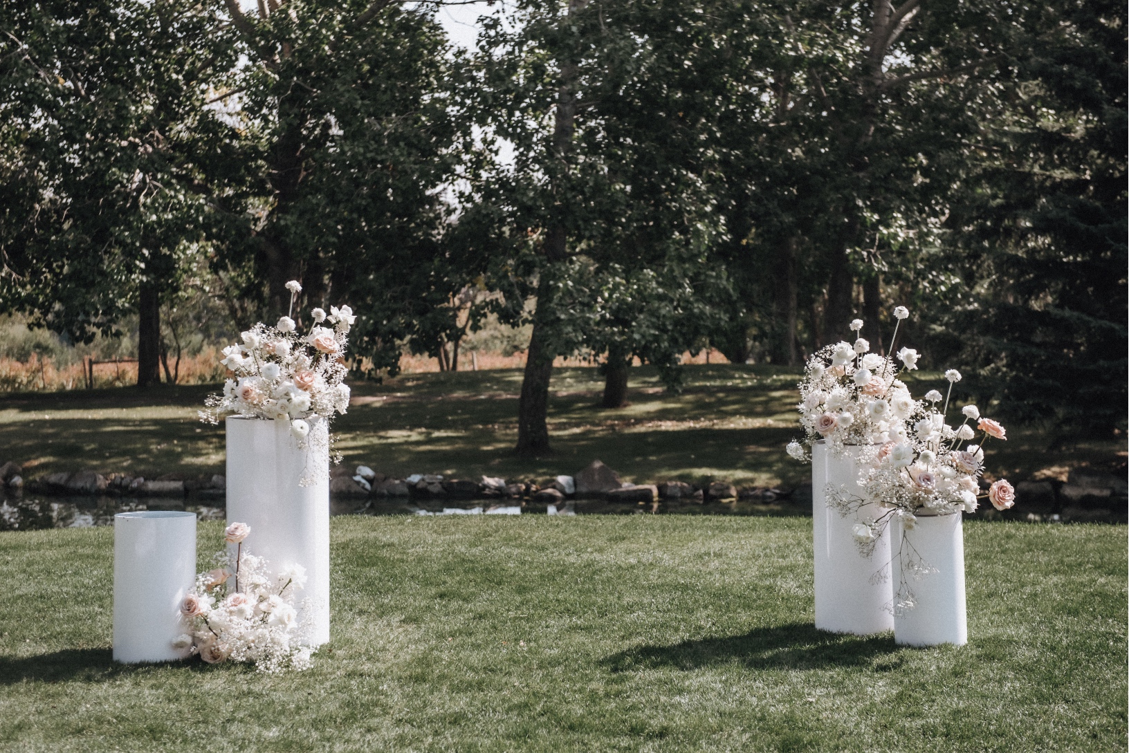 Pillar Arrangements Contemporary white pillars with arrangements of Playa Blanca roses, Quicksand roses, ivory spray roses, ranunculus, scabiosa, baby's breath and bleached ruscus with Hilltop's pond in the background.