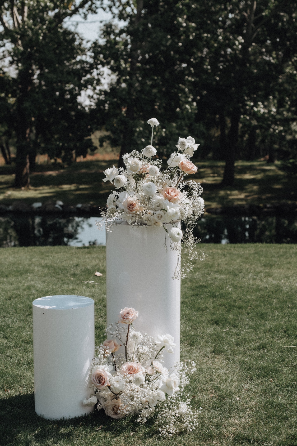 Pillar Set Contemporary white pillars with arrangements of Playa Blanca roses, Quicksand roses, ivory spray roses, ranunculus, scabiosa, baby's breath and bleached ruscus with Hilltop's pond in the background.